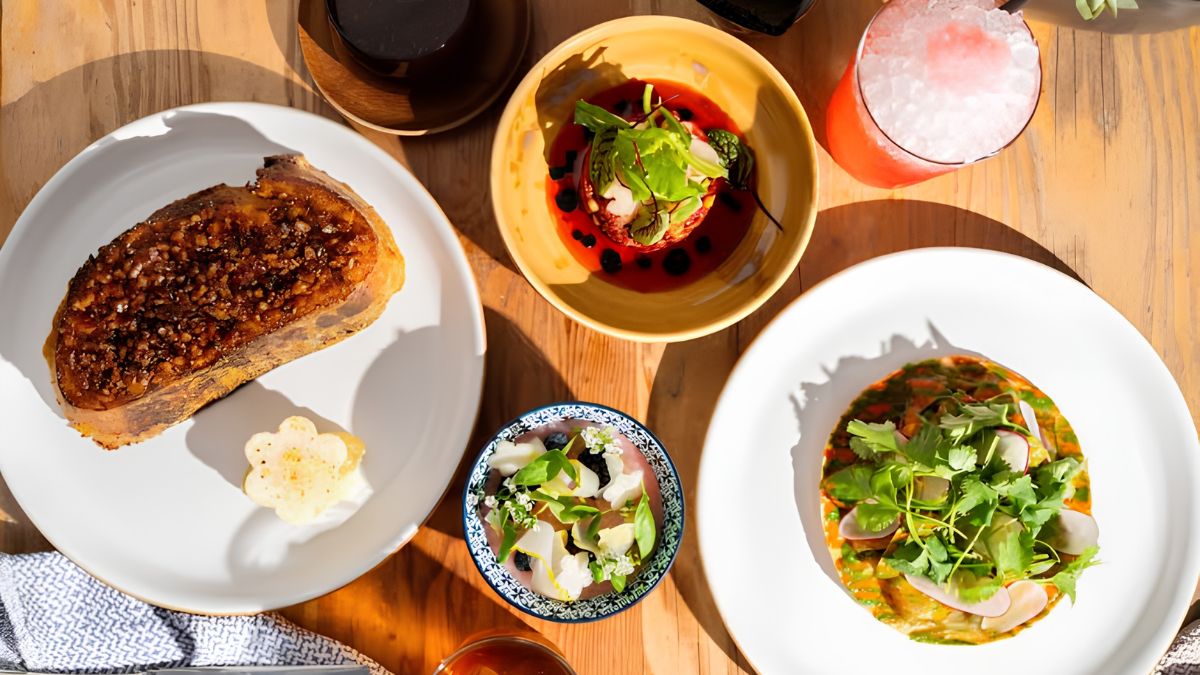 An overhead flatlay view of a wooden table spread with gourmet dishes, plates, and a refreshing cocktail.