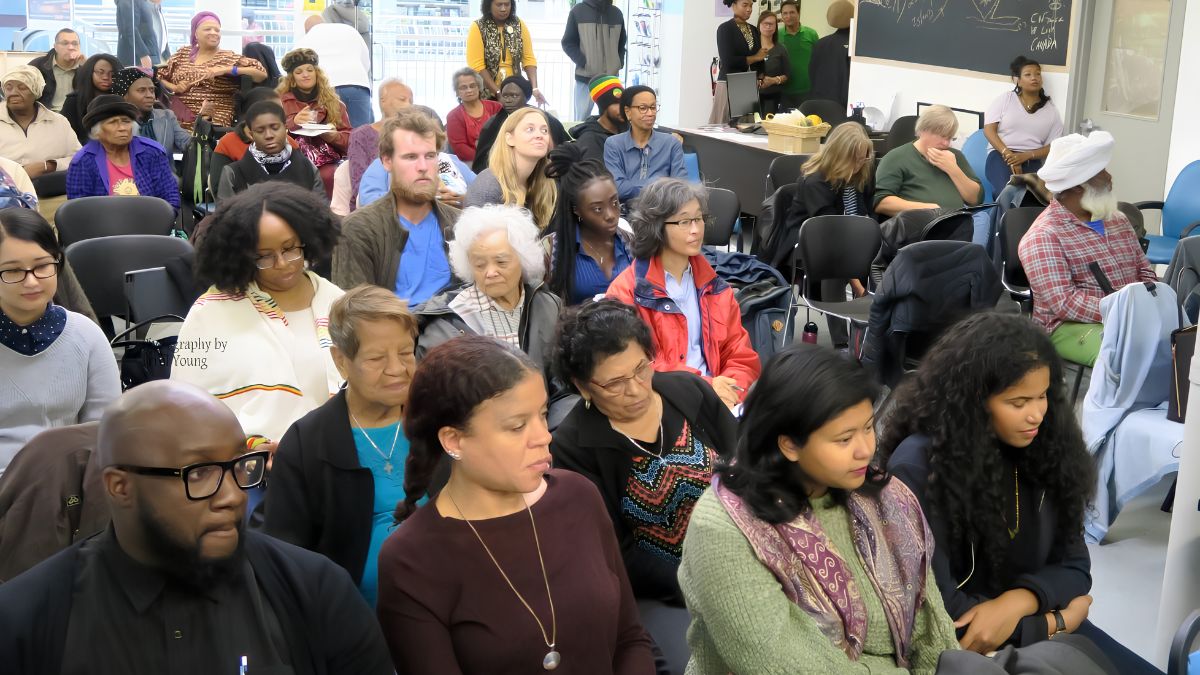 A diverse crowd of attentive people of various ages and backgrounds seated in rows, listening intently during a community meeting or public seminar.